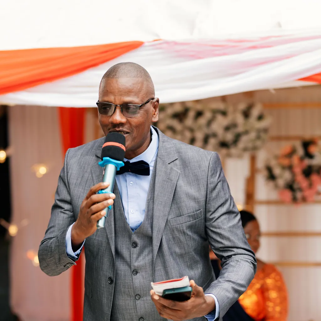 Event host in a gray suit and bow tie speaking into a handheld microphone at a festive event
