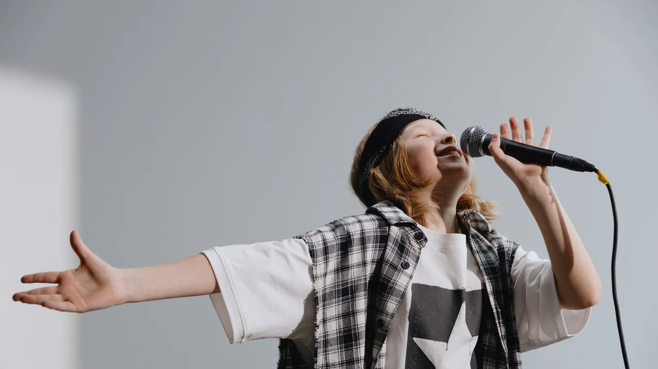 Kid in a plaid shirt belting into a microphone with one arm stretched out