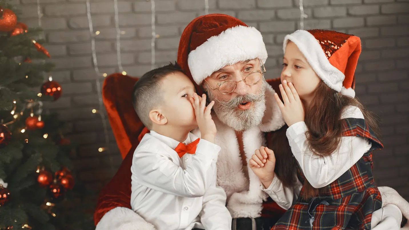 Two children whispering their Christmas wishes to Santa Claus beside a decorated tree