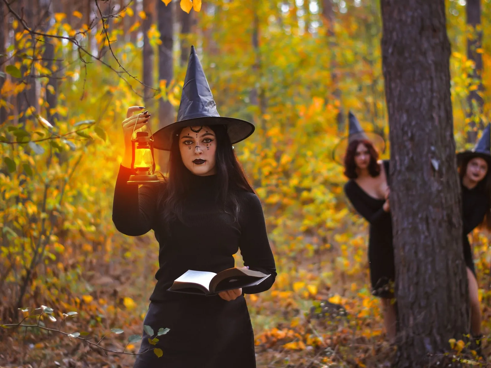 Woman dressed as a witch holding a lantern in an autumn forest while two more witches peek from the trees