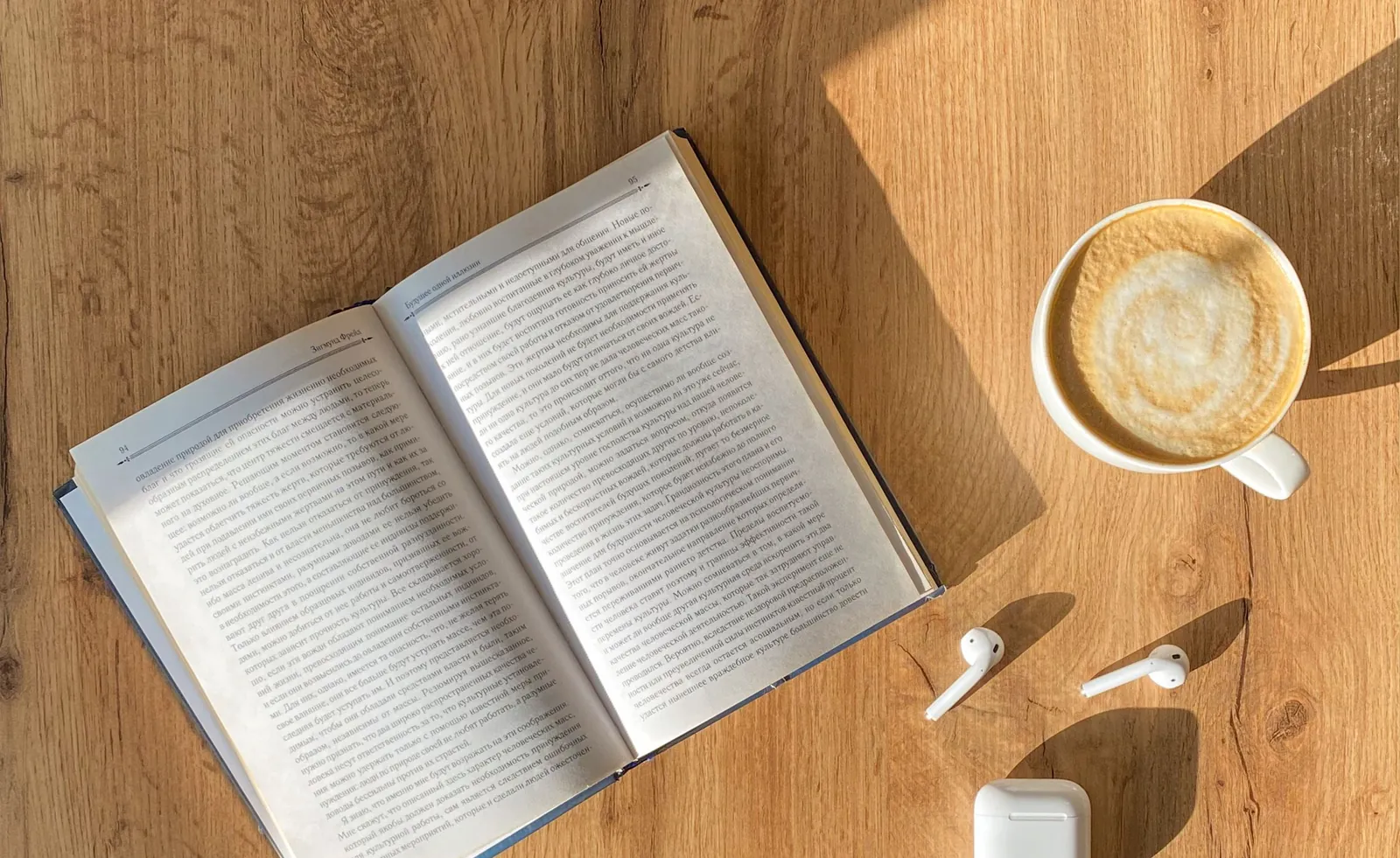 Open book, a latte and wireless earbuds on a sunny wooden table