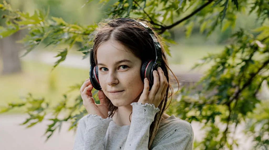 Girl holding her over-ear headphones and listening closely under green trees