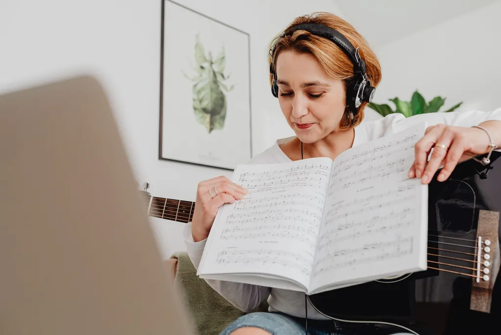 Woman in headphones taking a voice range test on her laptop, sheet music in hand and a guitar on her lap