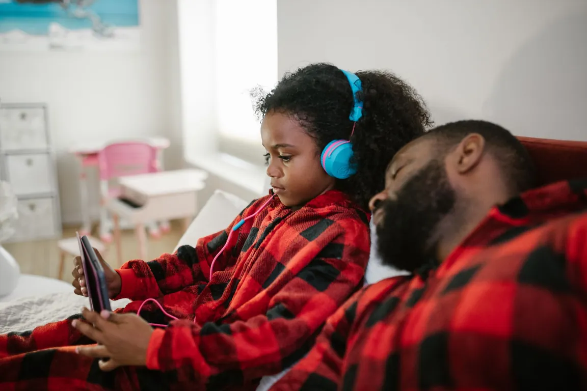 Girl in headphones watching a tablet in bed next to her dad, both in red plaid pajamas