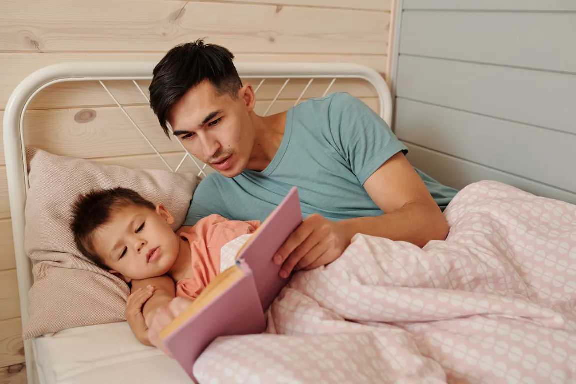 Dad reading a picture book to his sleepy little daughter in bed