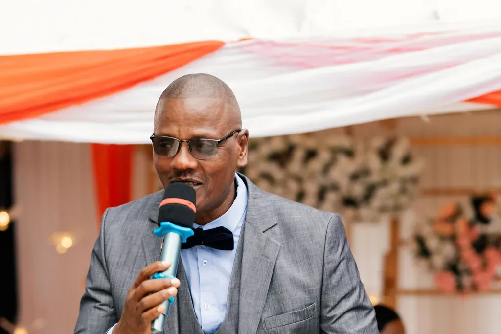 Event host in a gray suit and bow tie announcing into a handheld microphone