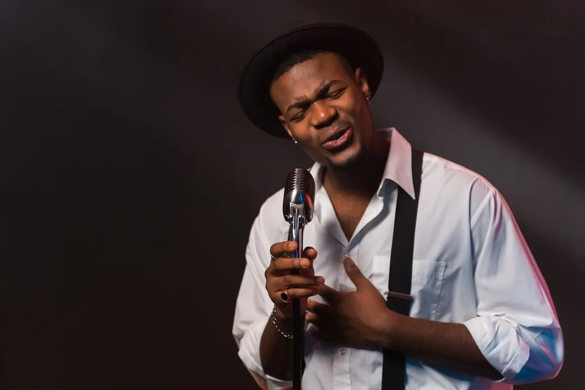 Singer in a hat and suspenders crooning into a vintage microphone on a dark stage