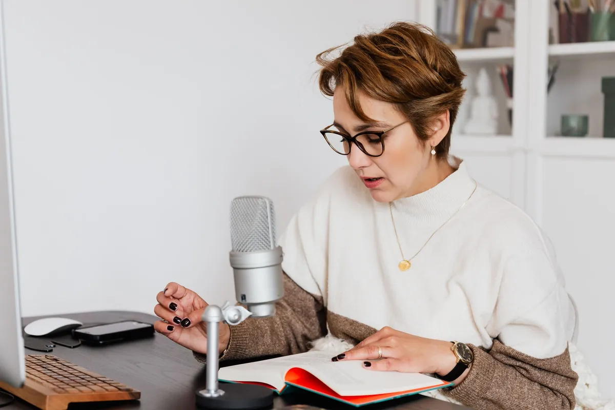 Woman in glasses reading a book aloud into a desk microphone, recording an audiobook
