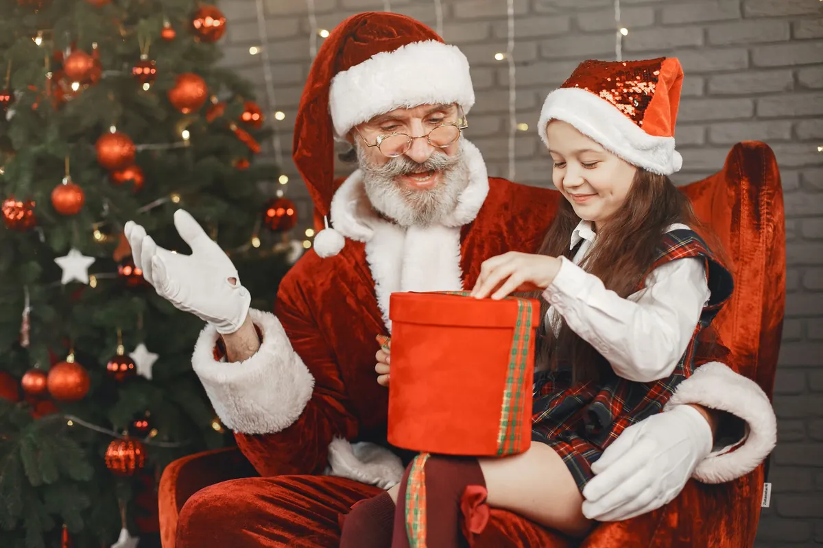 Santa Claus and a little girl laughing over a red gift box beside the Christmas tree