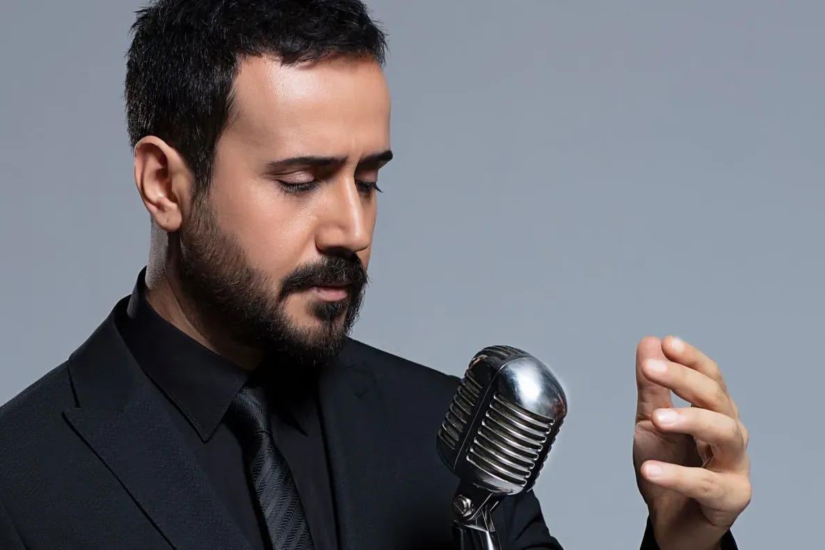 Bearded man in a black suit holding a vintage microphone stand