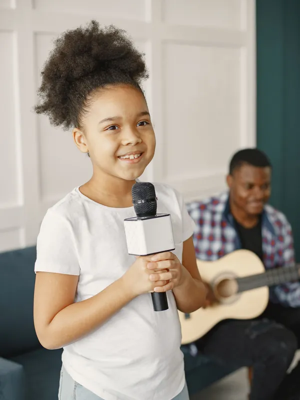 Smiling girl holding a microphone at home while her dad plays the guitar behind her