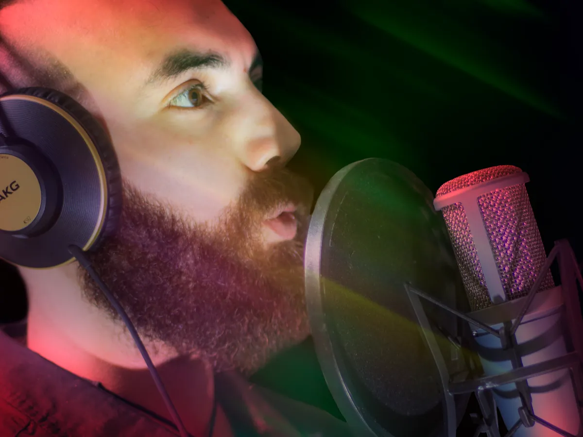 Bearded singer in headphones holding a note close to a studio microphone under colorful lights