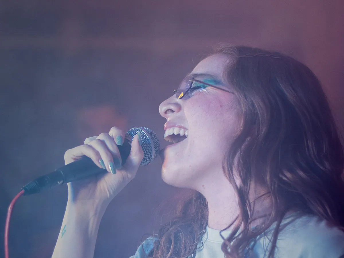 Singer with glasses belting into a handheld microphone on a stage lit in purple