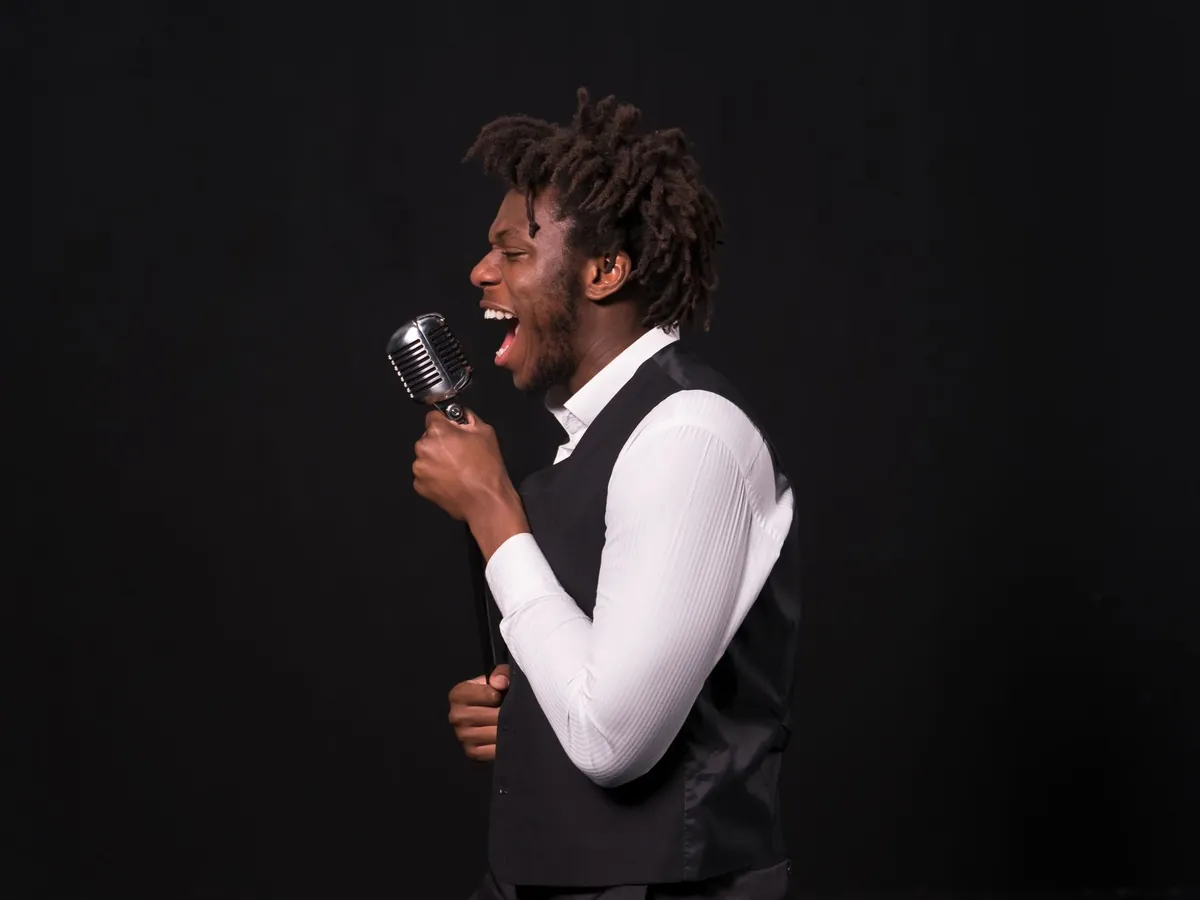 Singer in a white shirt and vest singing into a vintage microphone against a black background
