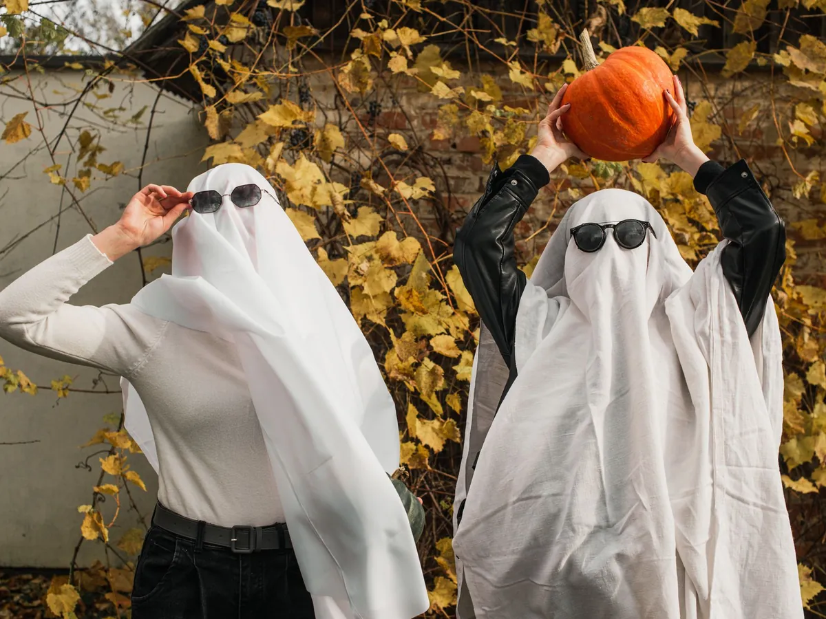 Friends in ghost sheets and sunglasses holding up a pumpkin among autumn leaves