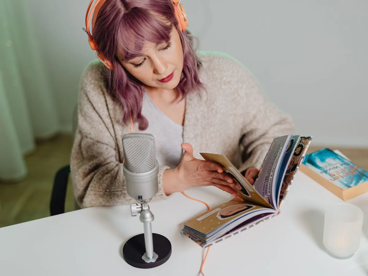 Woman with pink hair reading a book aloud at a desk microphone