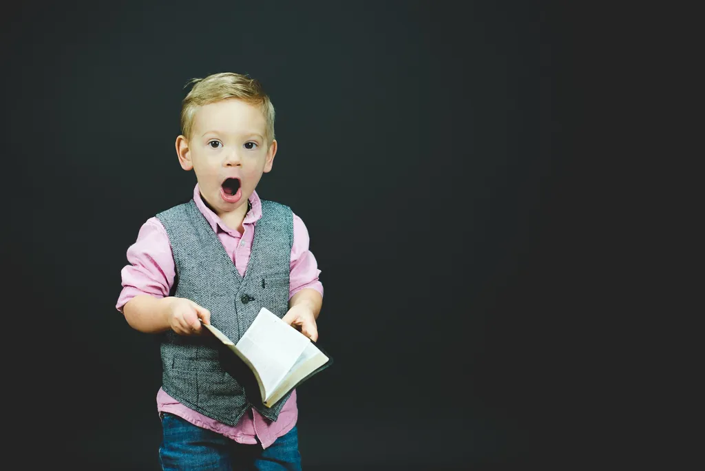 A little boy in a gray vest reading aloud from an open book, mouth wide with excitement