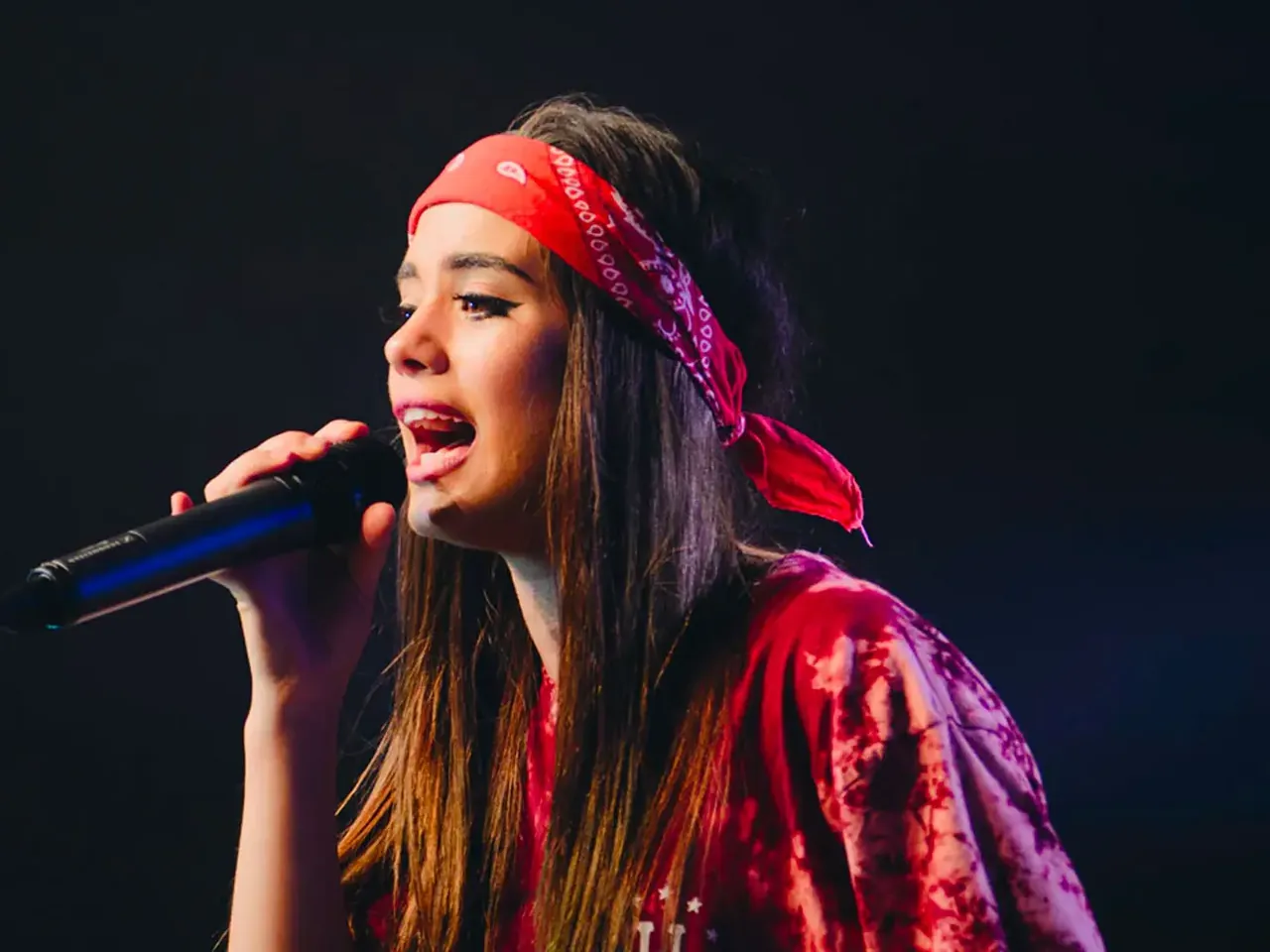 Young woman in a red bandana singing into a handheld microphone on a dark stage, showing off her mezzo-soprano range
